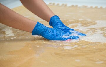 Two hands wearing blue nitrile gloves press down on a pink sponge that is placed on top of a textile. White foam is floating in the shallow water bath that the textile is resting in.